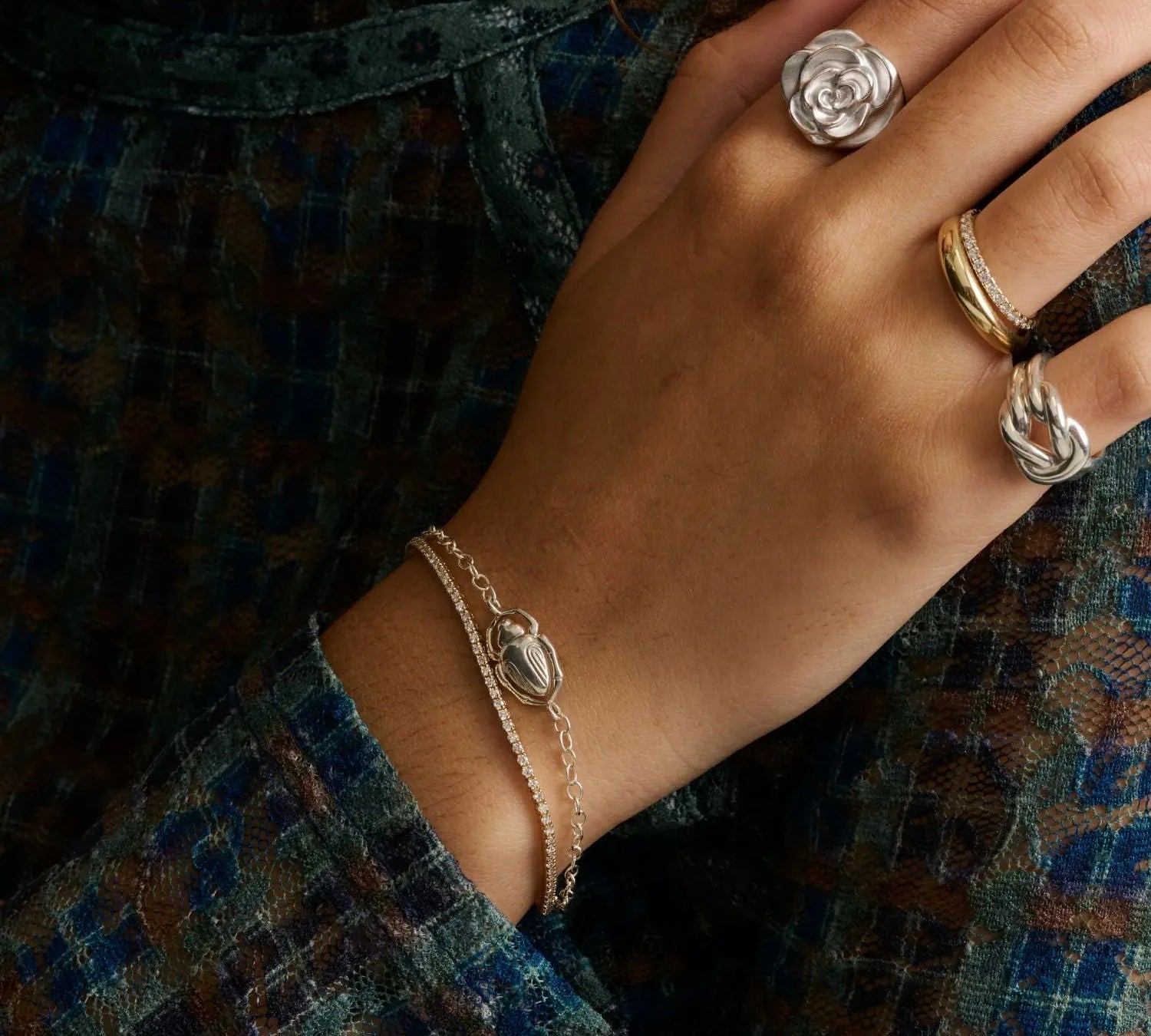A close-up of a hand wearing multiple rings and a double-strand chain bracelet, styled with a textured blue and brown plaid garment.