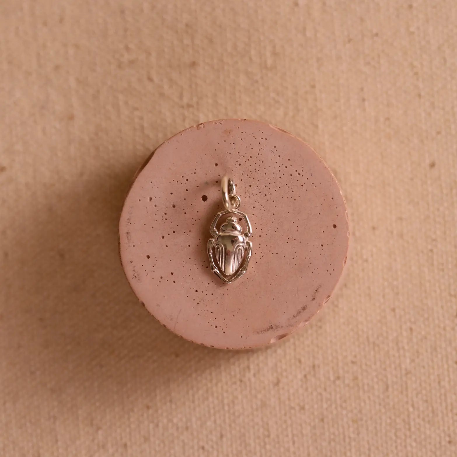 Silver scarab charm on a pink ceramic circular pedestal against a neutral fabric background