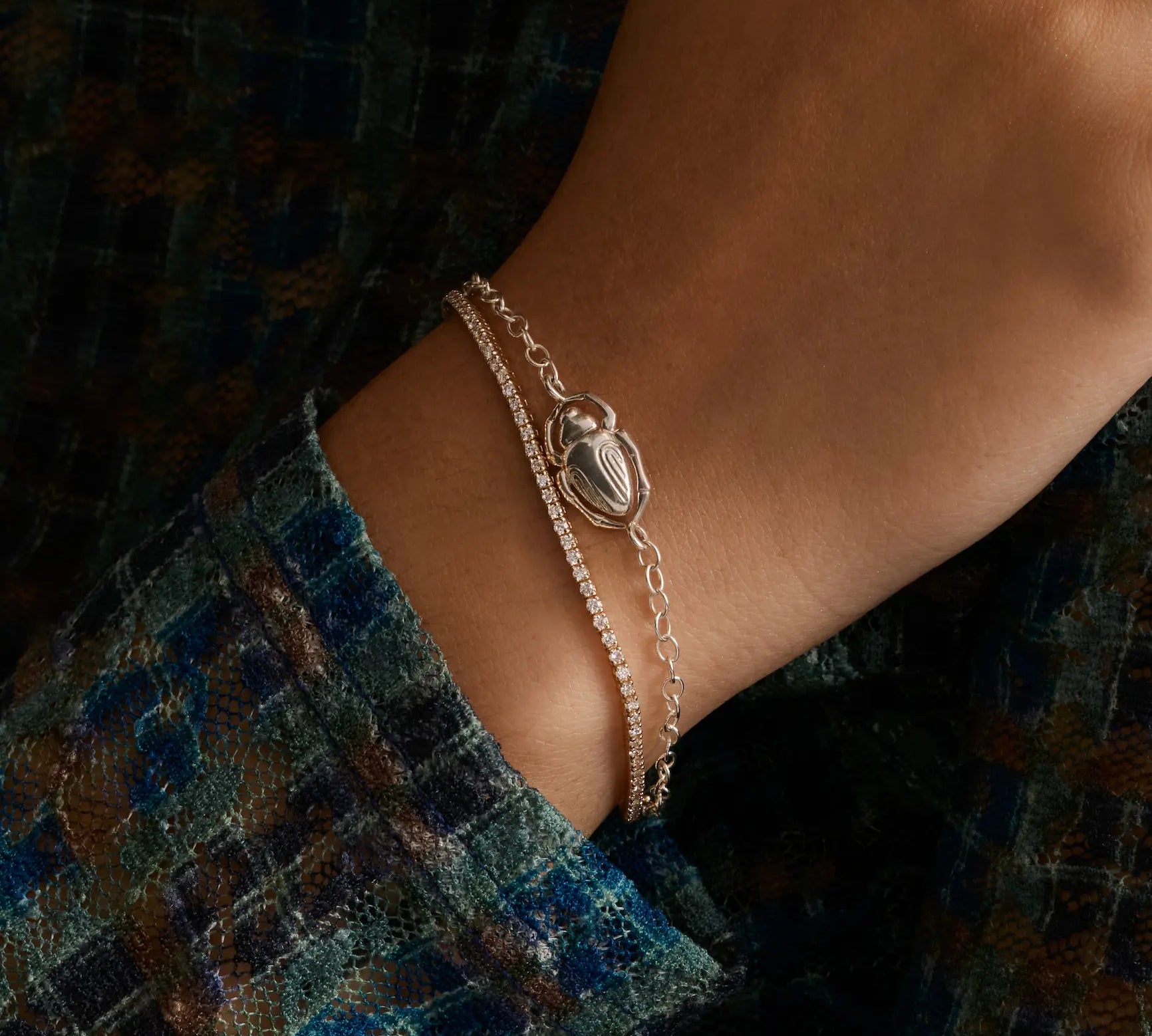 close up of woman's wrist with a dainty diamond tennis bracelet and a silver bracelet with a scarab 