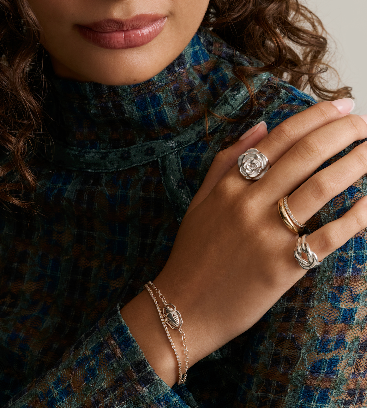 A close-up lifestyle shot of a model wearing a silver rose ring, a gold band, and a silver leaf ring on her fingers, paired with a textured blue and brown tweed turtleneck.