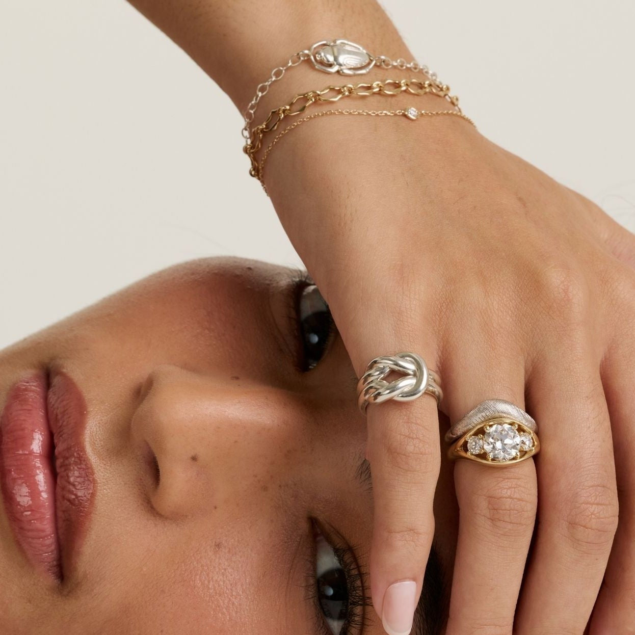 A close-up lifestyle shot of a model wearing gold and silver rings, delicate gold bracelets, and a small gold hoop earring. The model is lying down, looking towards the camera, with her hand resting near her face against a clean, light beige background.