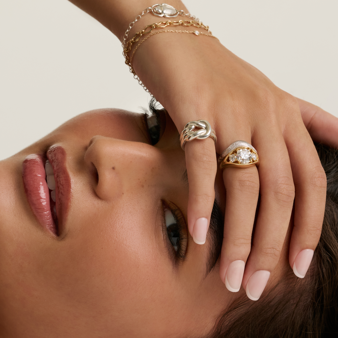 A close-up lifestyle shot of a model wearing multiple gold rings, including a prominent gold ring with a white stone, along with matching gold bracelets and earrings.
