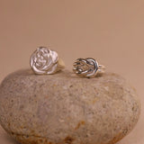 Two silver rings, one rose-shaped and one knot-shaped, on a stone surface with a beige background.