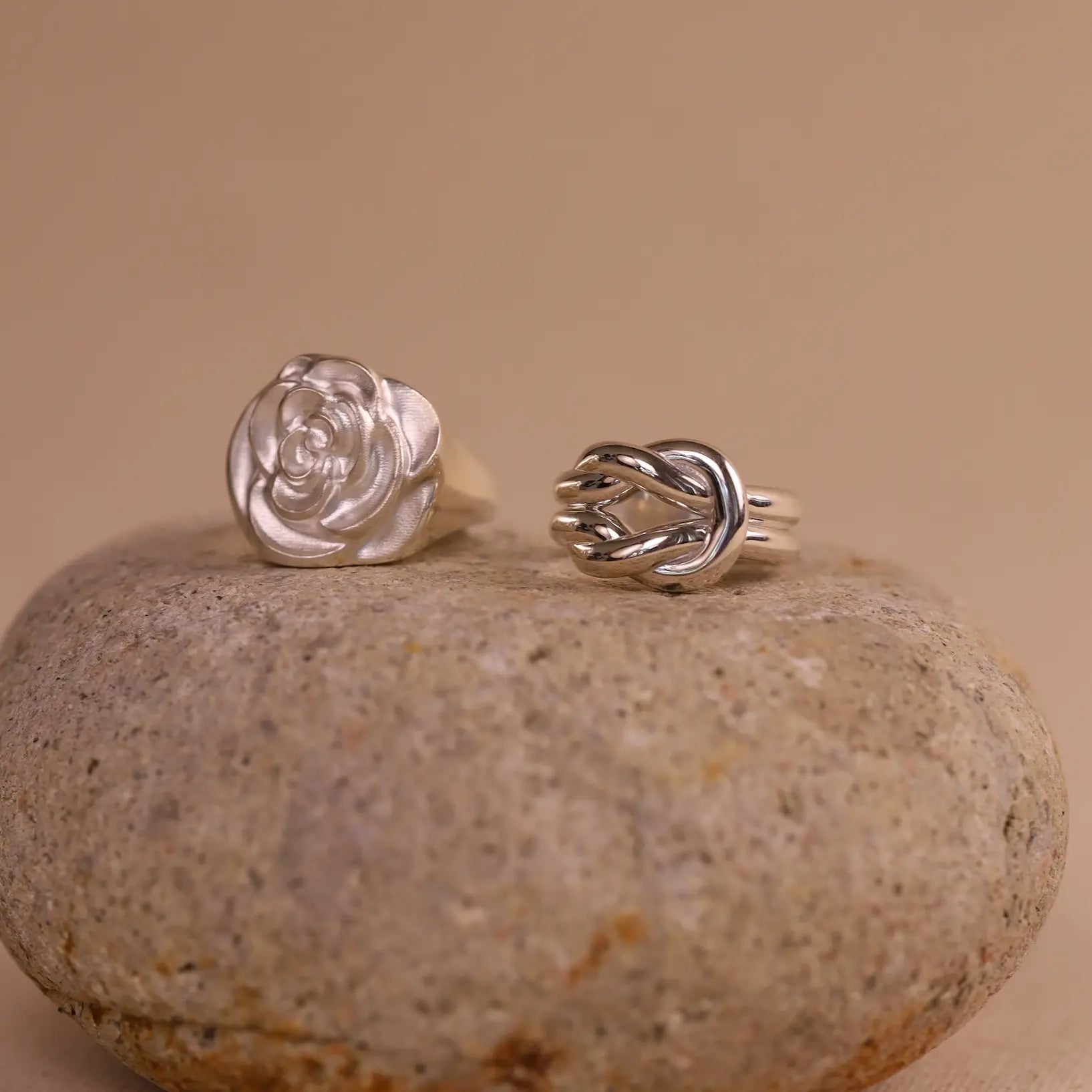 Two silver rings, one rose-shaped and one knot-shaped, on a stone surface with a beige background.