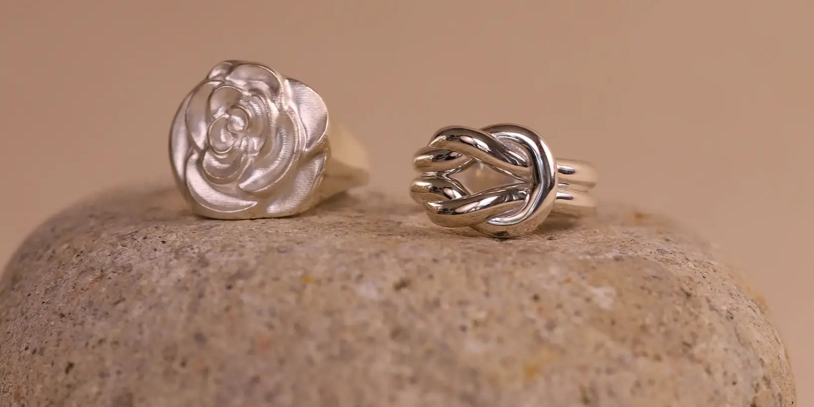 Two silver rings, one rose-shaped and one knot-shaped, on a stone surface with a beige background.