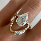 A close-up of a gold ring stack worn on a finger. The stack features a yellow gold band with a large pear-cut clear gemstone and a smaller curved gold band with small round clear gemstones.