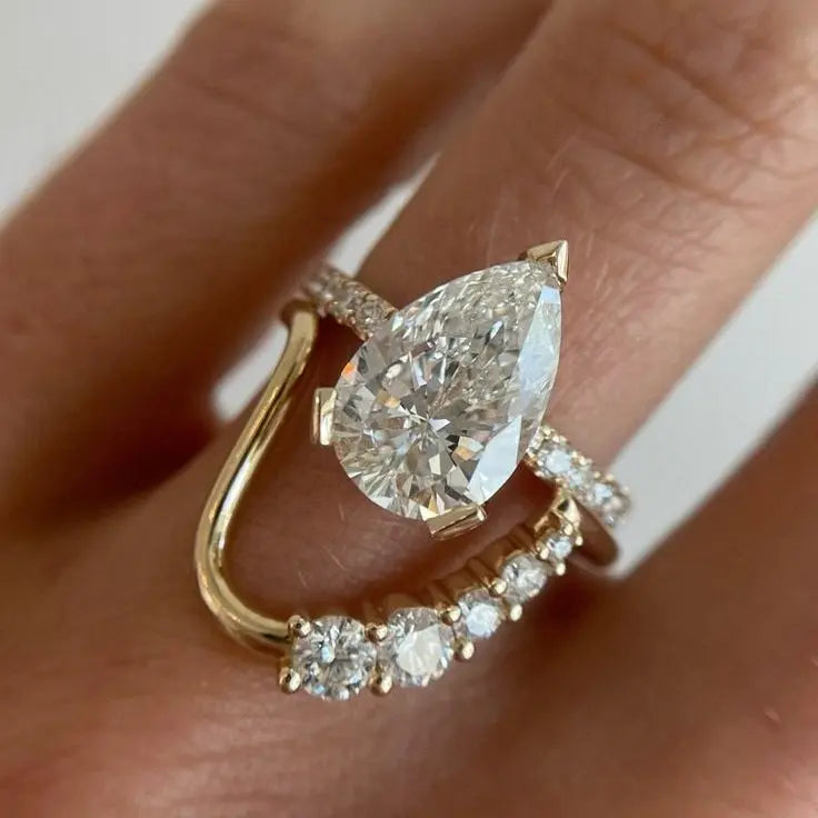 A close-up of a gold ring stack worn on a finger. The stack features a yellow gold band with a large pear-cut clear gemstone and a smaller curved gold band with small round clear gemstones.