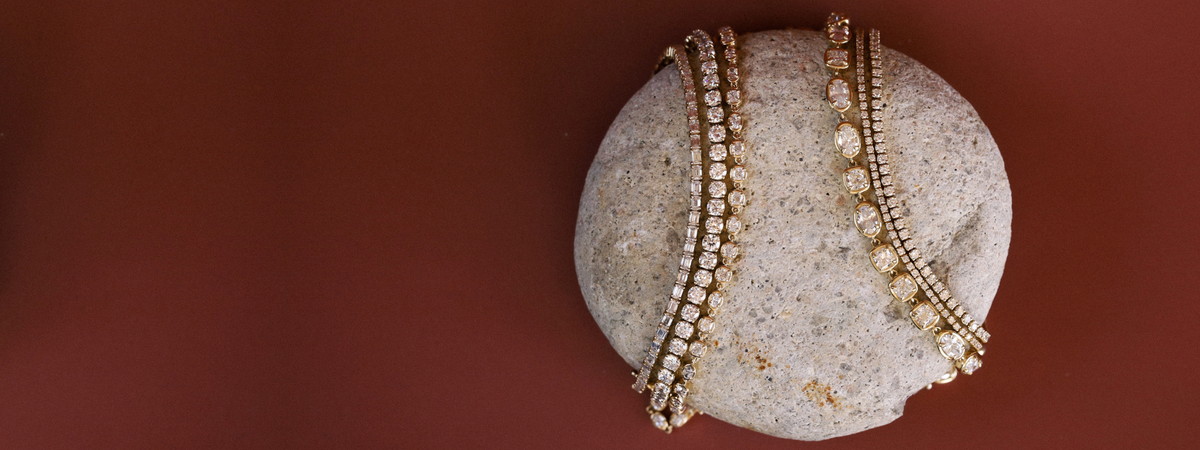 Six tennis bracelets draped over a round beige stone, set against a solid dark reddish-brown background.
