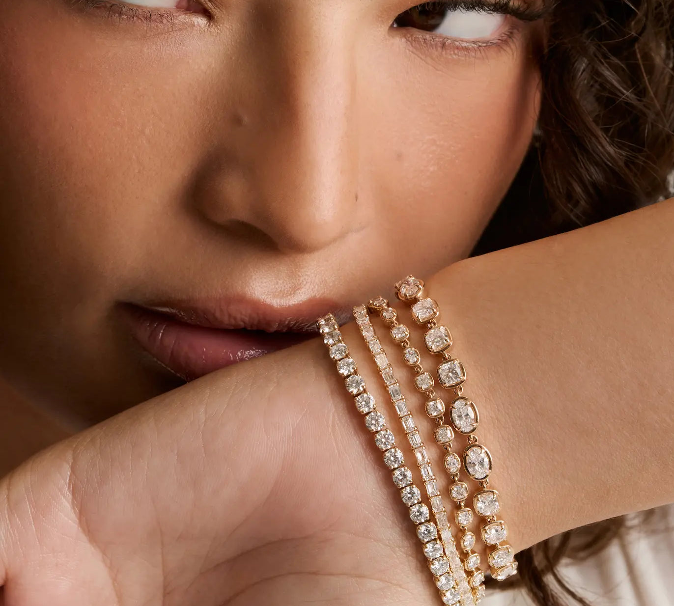 A close-up of a model's wrist wearing three delicate rose gold tennis bracelets adorned with sparkling clear gemstones, set against a soft-focus background.