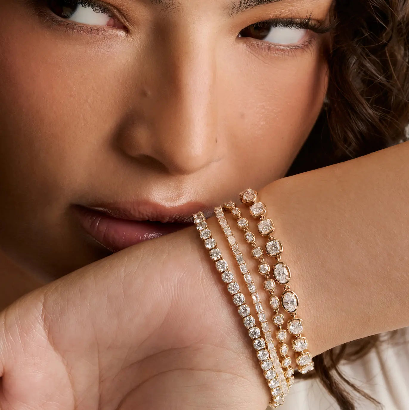 A close-up of a model's wrist wearing three delicate rose gold tennis bracelets adorned with sparkling clear gemstones, set against a soft-focus background.