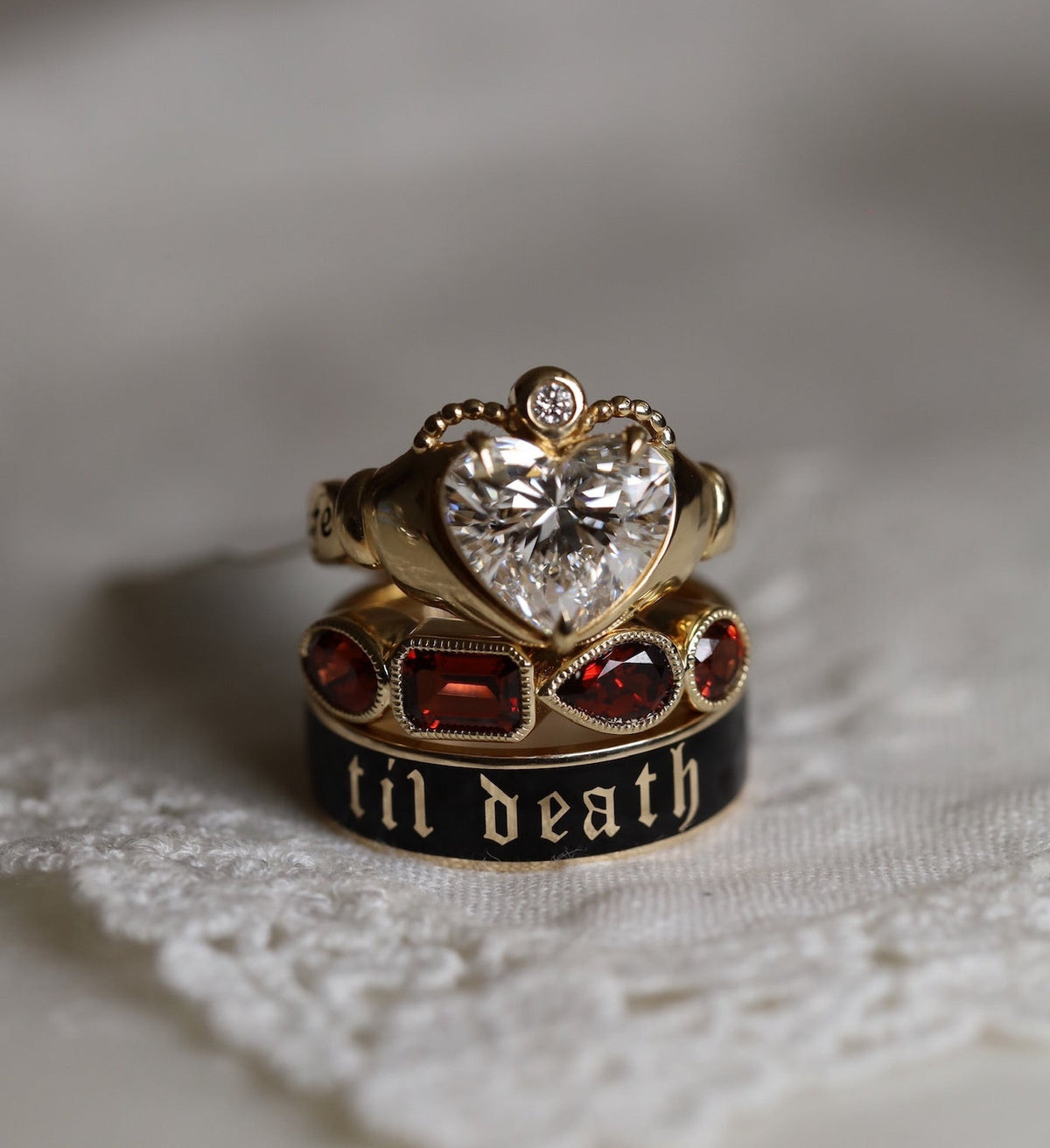 A gold ring with a heart-shaped clear gemstone and a smaller ring featuring red stones and the gothic text 'til death' on a black band, displayed on a white textured fabric.