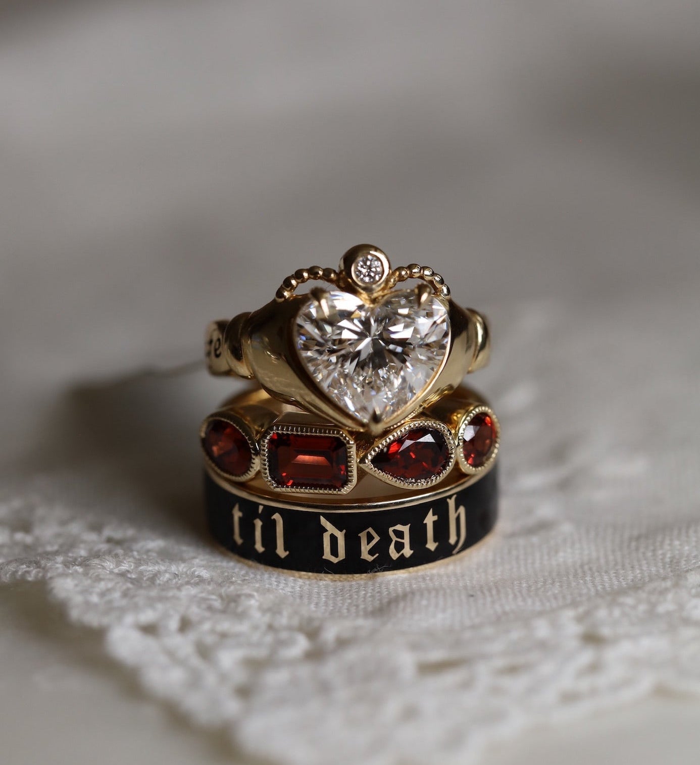 A gold ring with a heart-shaped clear gemstone and a smaller ring featuring red stones and the gothic text 'til death' on a black band, displayed on a white textured fabric.