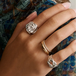 Close-up of hand wearing two chunky silver rings—one knot and one rose—and a gold dome band stacked with a thin pave diamond band