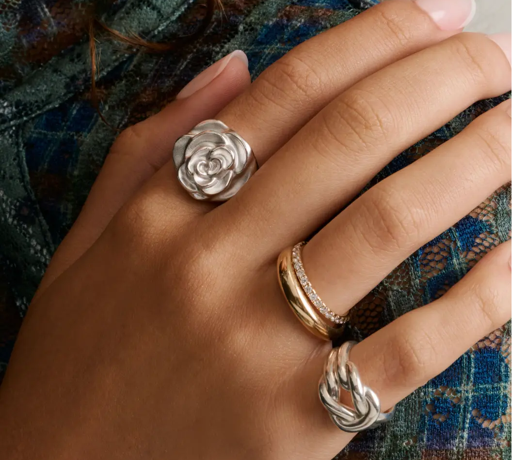 Close-up of hand wearing two chunky silver rings—one knot and one rose—and a gold dome band stacked with a thin pave diamond band