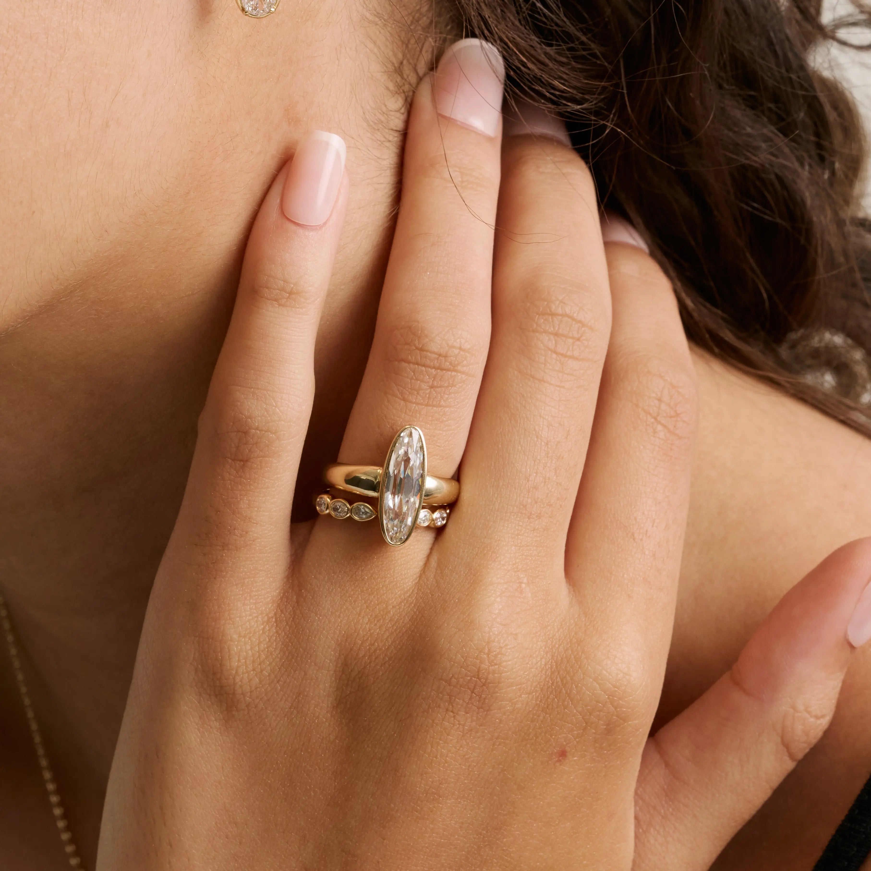 A close-up of a person's hand wearing a gold ring with a large oval clear gemstone and smaller side stones, shown against skin tones and dark hair.