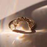 A gold ring with a row of small clear gemstones and one larger marquise-cut clear stone, photographed on a softly lit beige surface with dramatic light leaks and shadows.