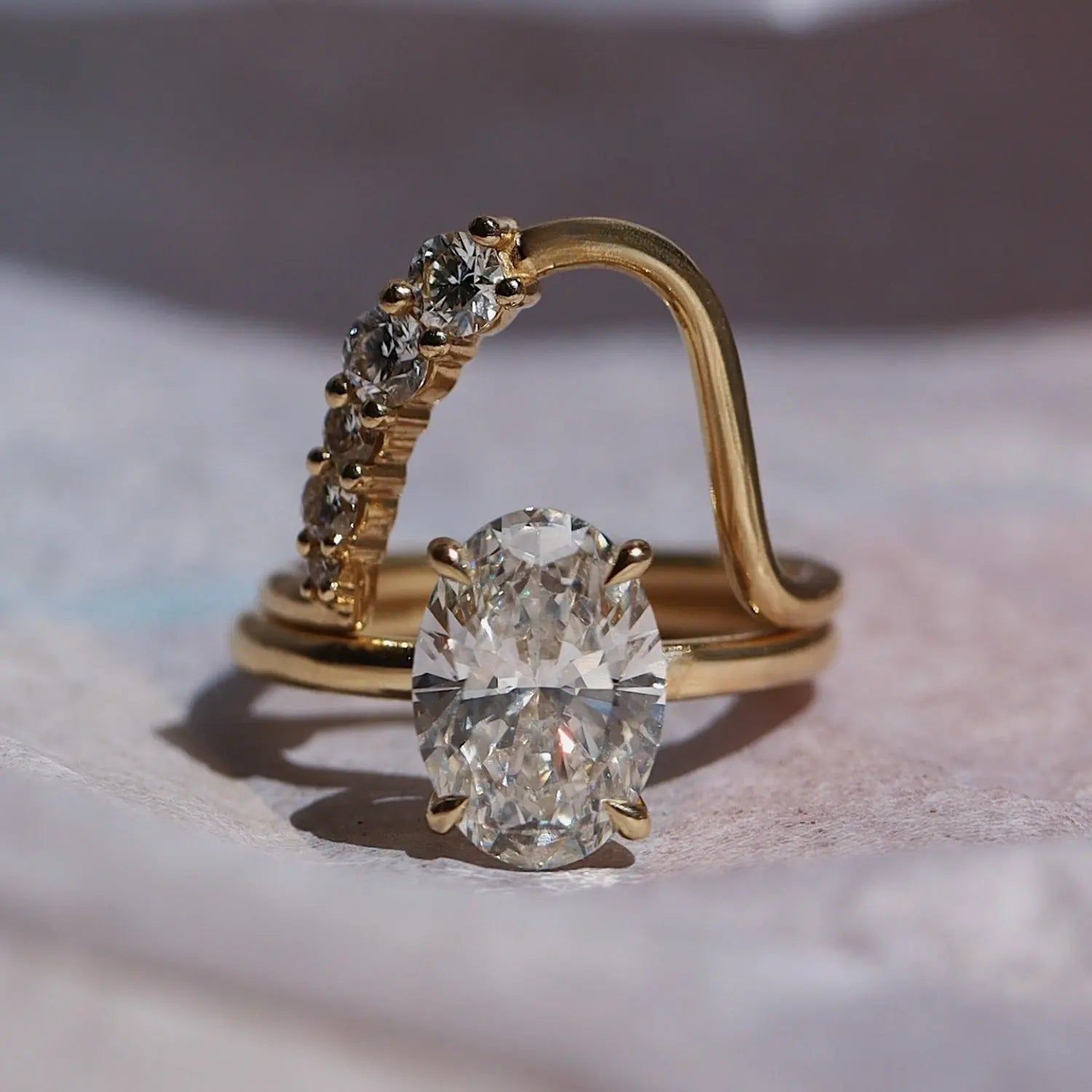 flatlay of yellow gold engagement ring with claw prongs and an oval diamond stacked with a waved asymmetrical band with round diamonds arching on one side, against a purplish pink background.