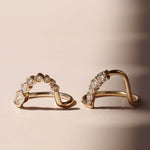 A pair of gold arched rings with rows of diamonds and round-cut stones, displayed on a warm beige surface with soft shadows.