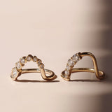 A pair of gold arched rings with rows of diamonds and round-cut stones, displayed on a warm beige surface with soft shadows.