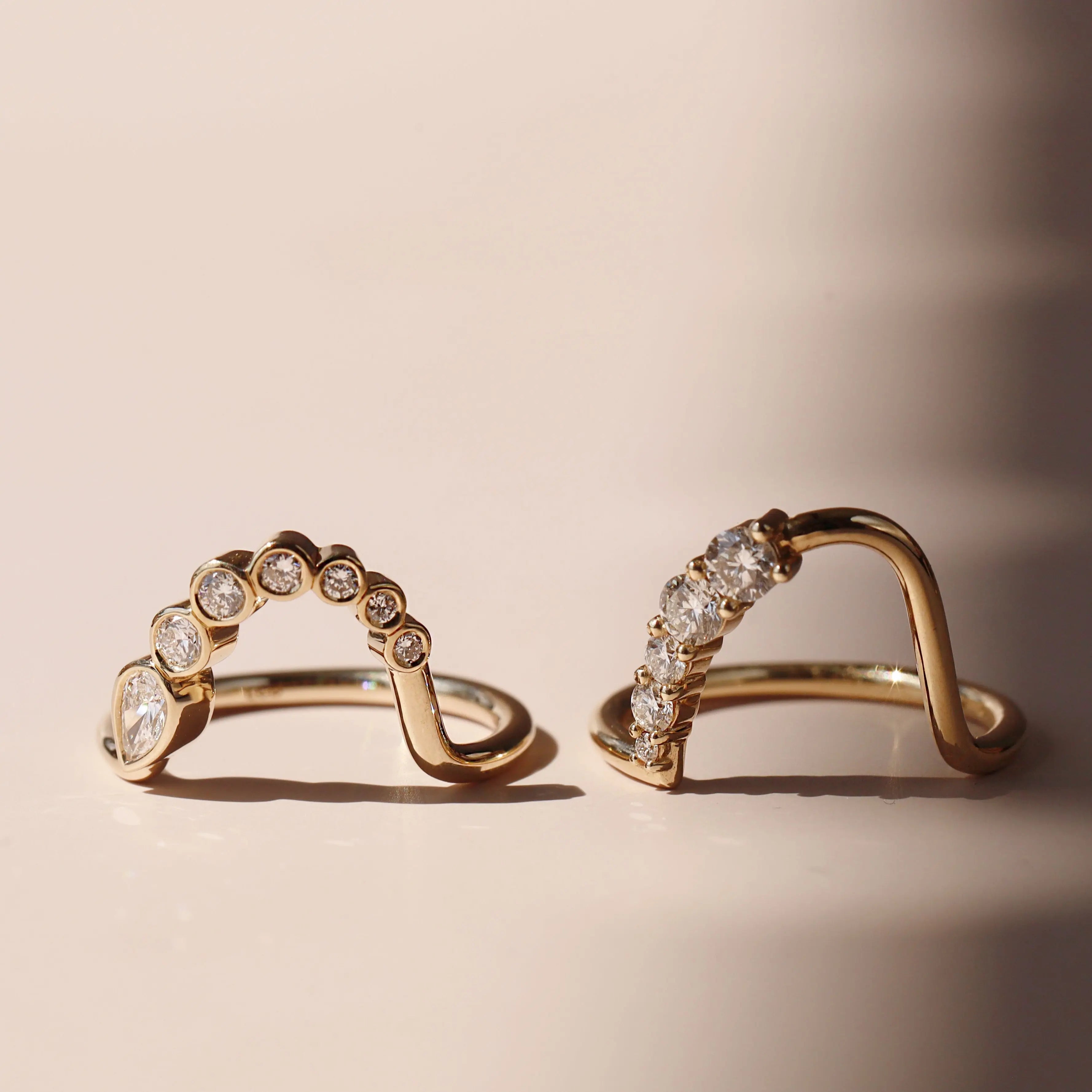 A pair of gold arched rings with rows of diamonds and round-cut stones, displayed on a warm beige surface with soft shadows.