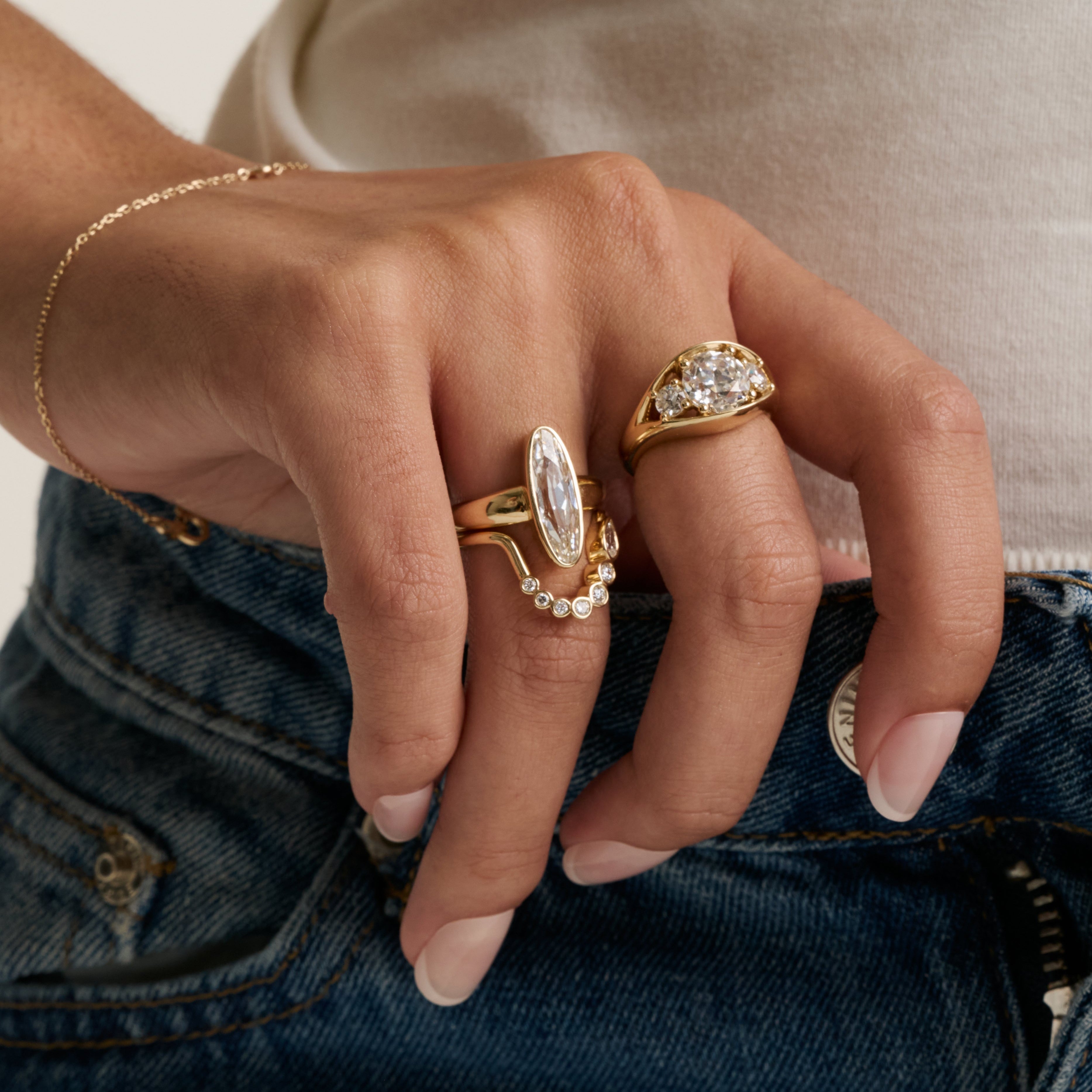 close up of woman's hand resting against top of denim wearing multiple yellow gold and diamond rings stacked together.